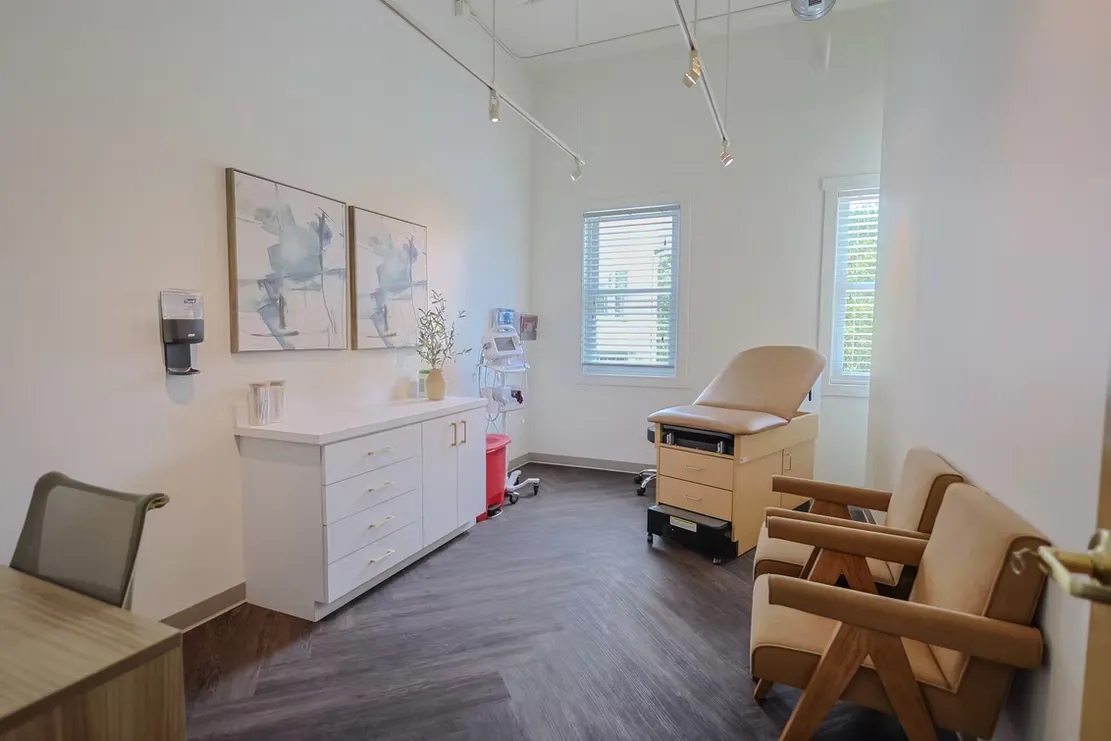 Medical examination room featuring a tan examination chair, white cabinetry with gold handles, medical equipment, abstract wall art, and large windows.