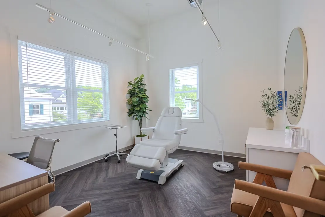 Bright, modern treatment room with a white treatment chair, large windows with blinds, a potted plant, and a clean minimalist interior.